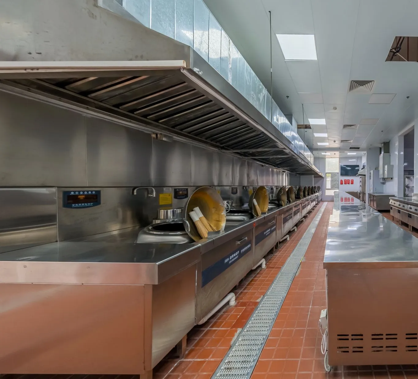 A long, empty stainless steel commercial kitchen with tiled floor and extraction hoods
