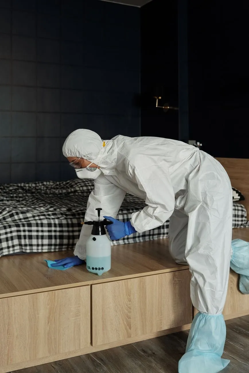 A technician in a white protective suit treating the frame of a bed with a hand sprayer