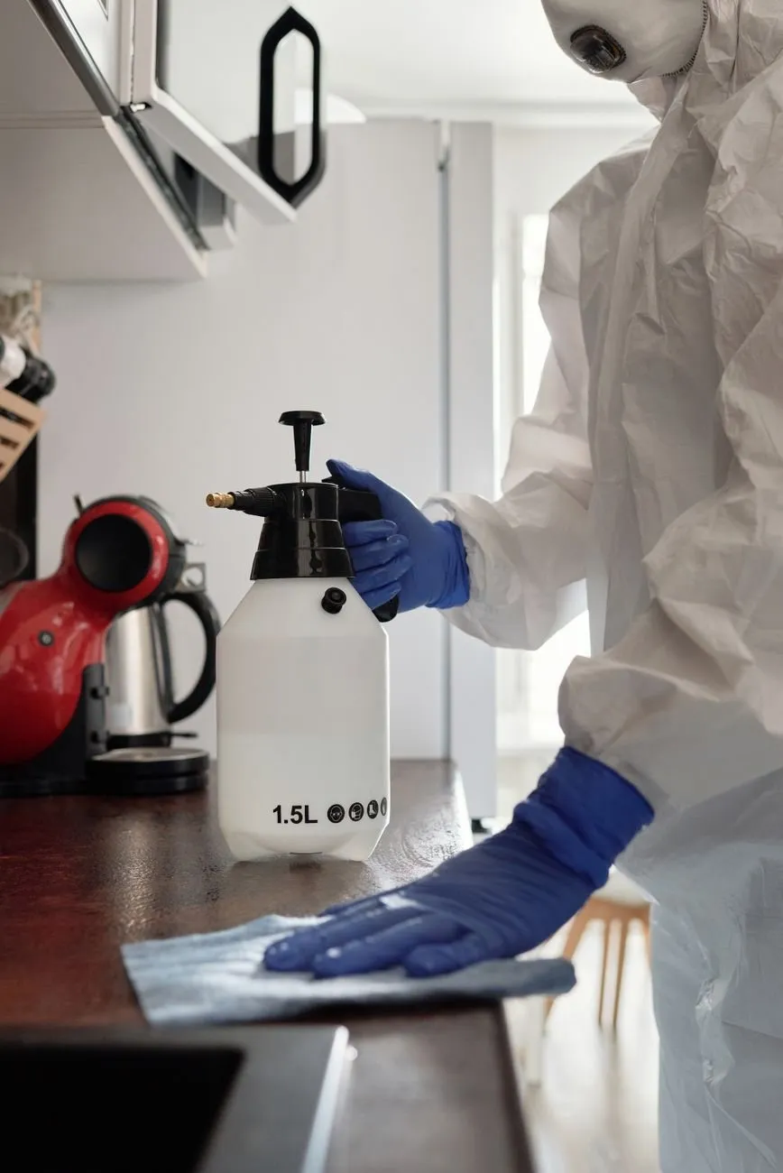 A technician in blue gloves and a white suit wiping a kitchen worktop beside a hand sprayer